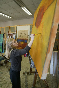 A ULM painting student wearning a gray shirt paints with a brush on a large canvas on an easel. 