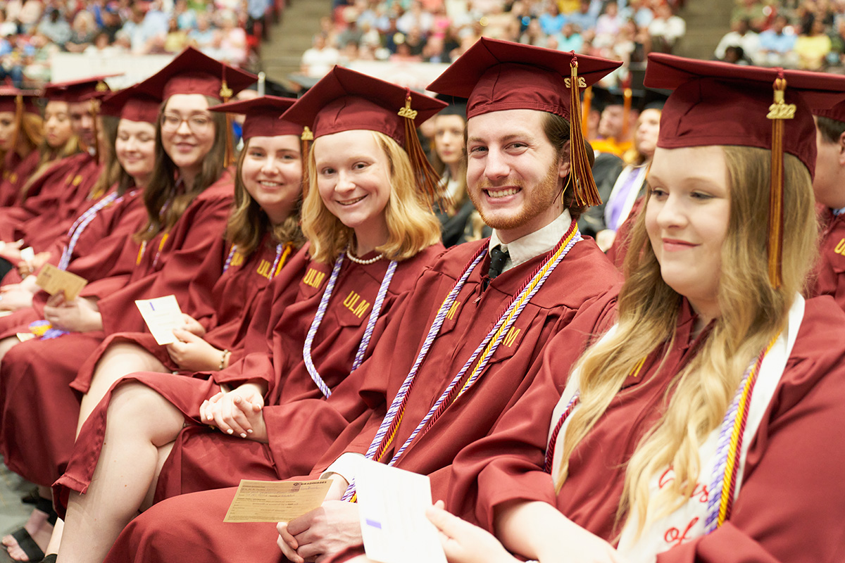 Students Graduating Row of students graduating seated, all wearing cap and gowns, audience in the background