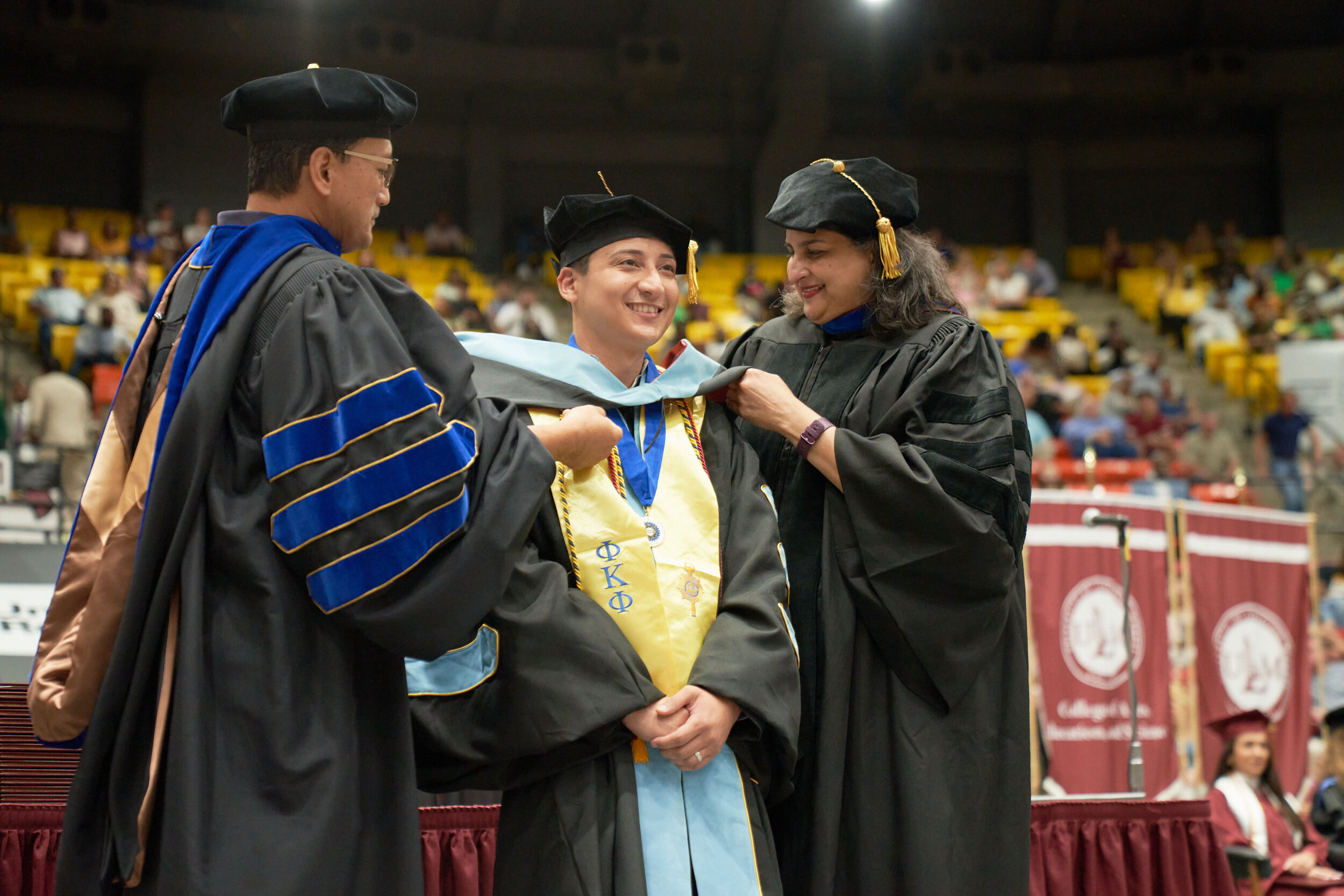 Hooding Doctoral hood being placed over head of male student by dean and professor, all are smiling, audience in background